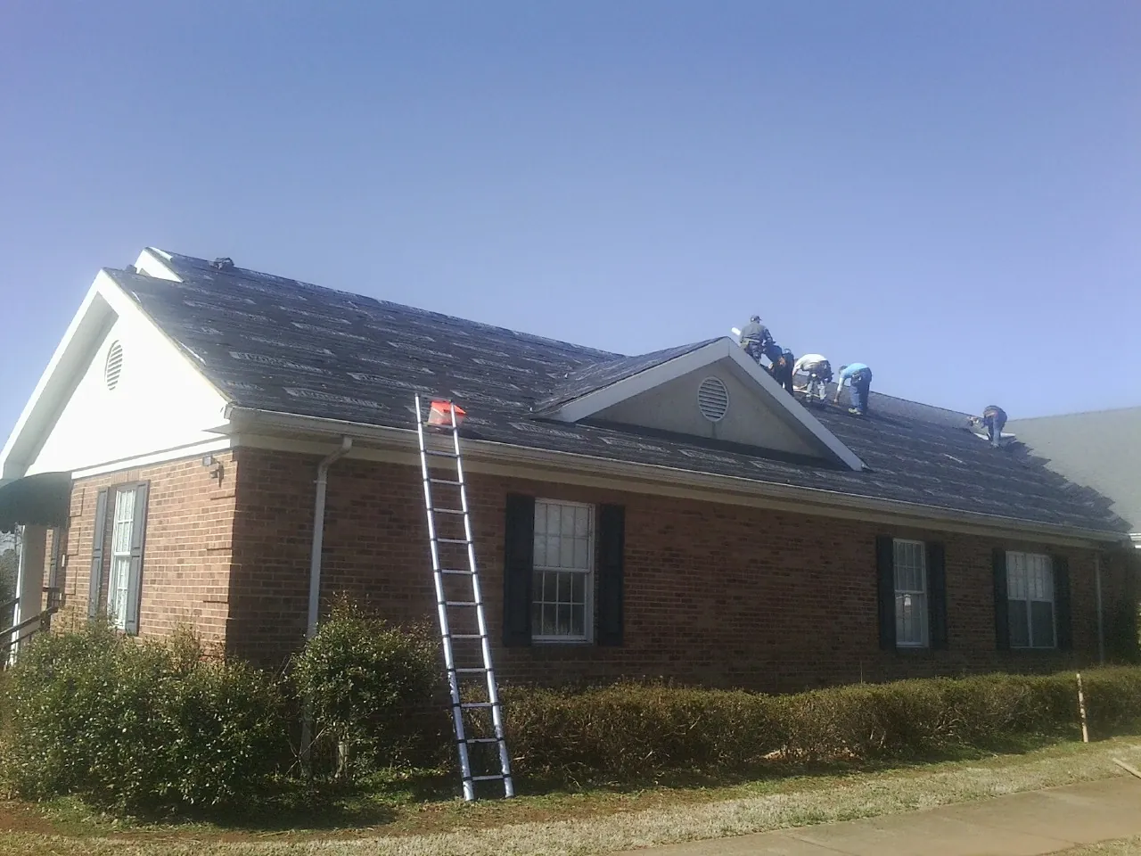 Crew tearing shingles off a brick ranch roof with a ladder set — Emergency Roof Repair work by our crew in Lansford