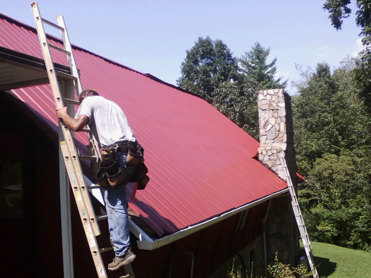 Roofer on a ladder beside a red metal roof and stone chimney — Soffit & Fascia Repair by Roofing Brothers in Lansford
